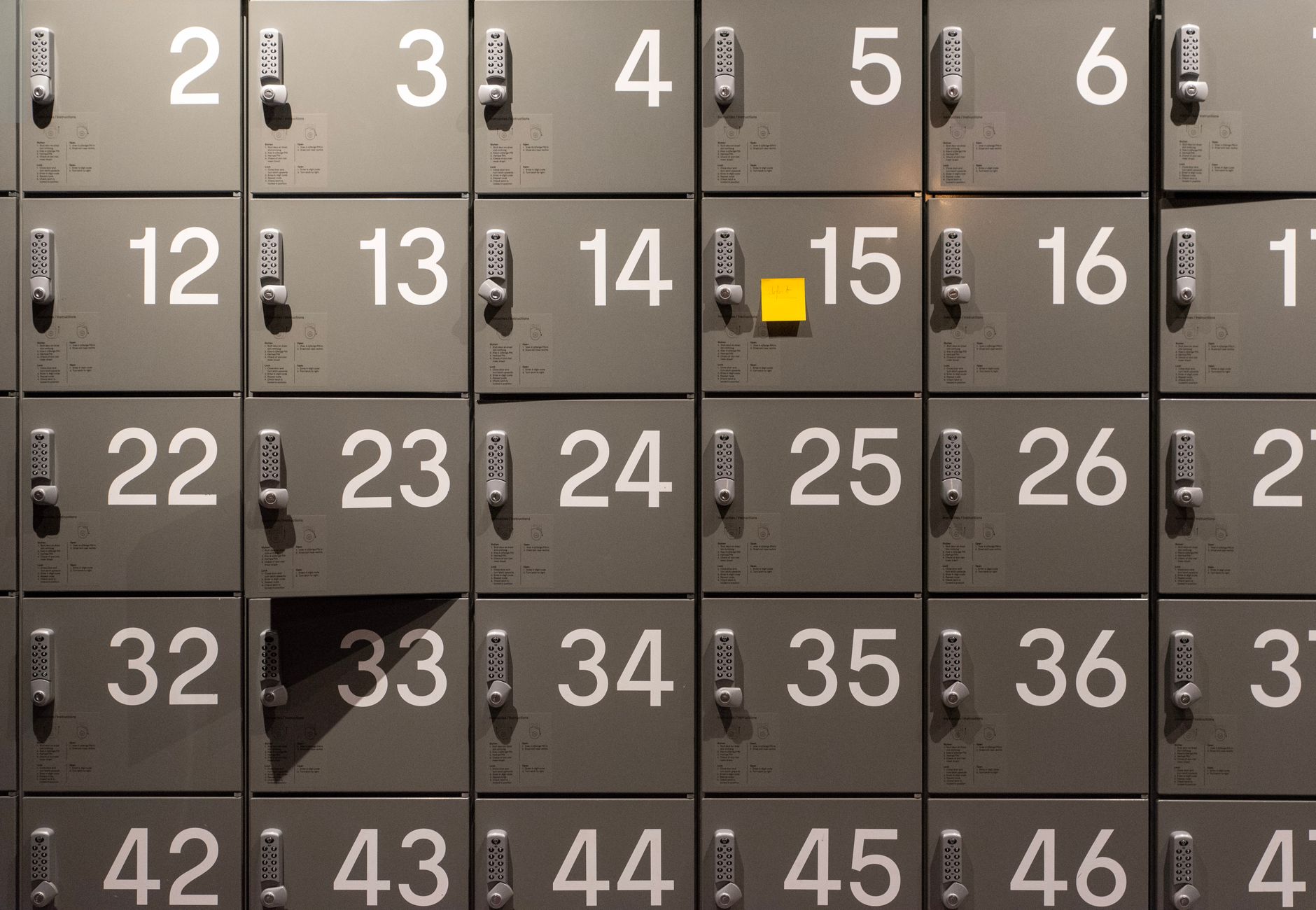 Gray lockers with electronic locks featuring numbers and a single yellow sticky note in the center.