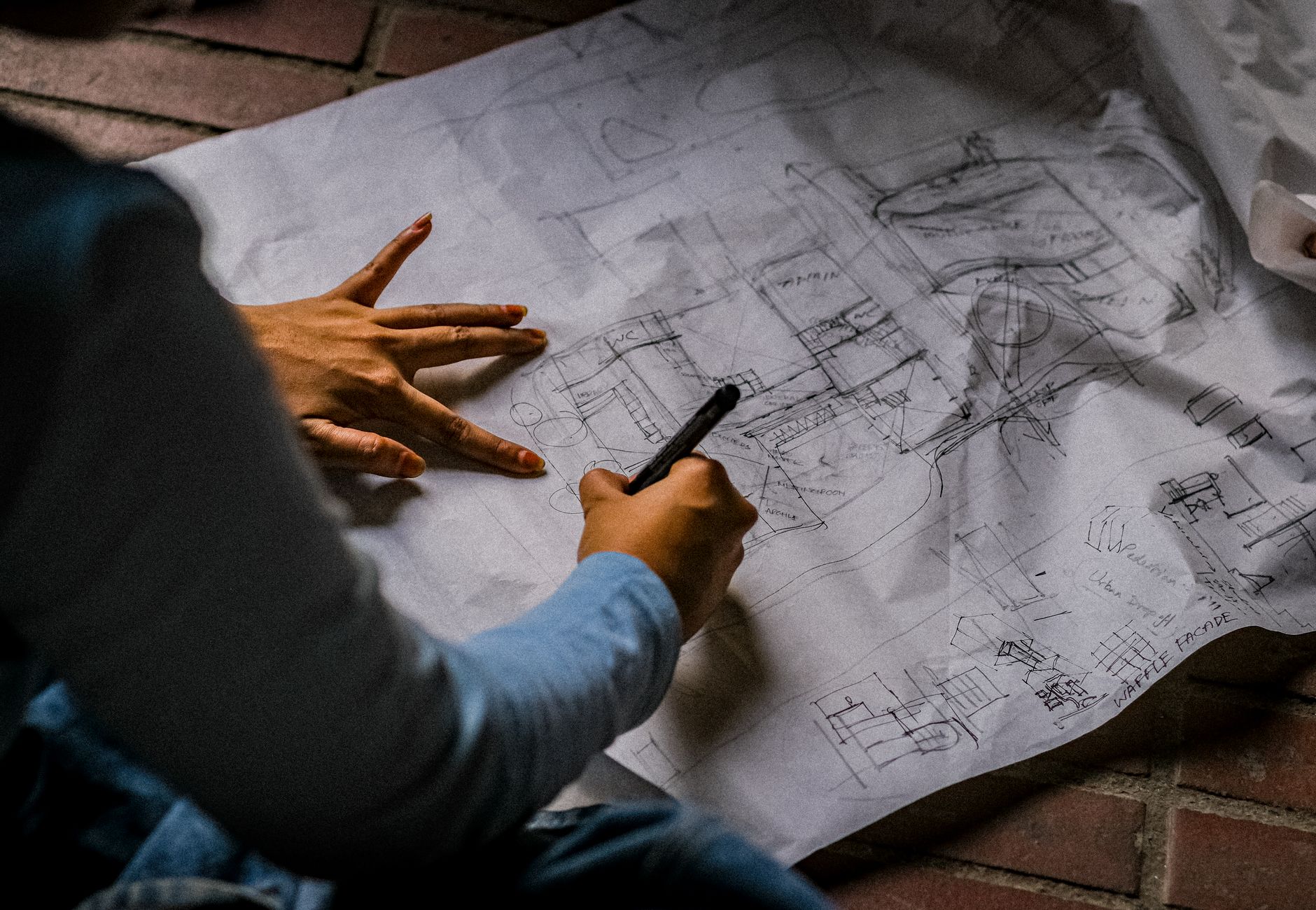 Person sketching architectural design on blueprint paper with pen on brick floor.